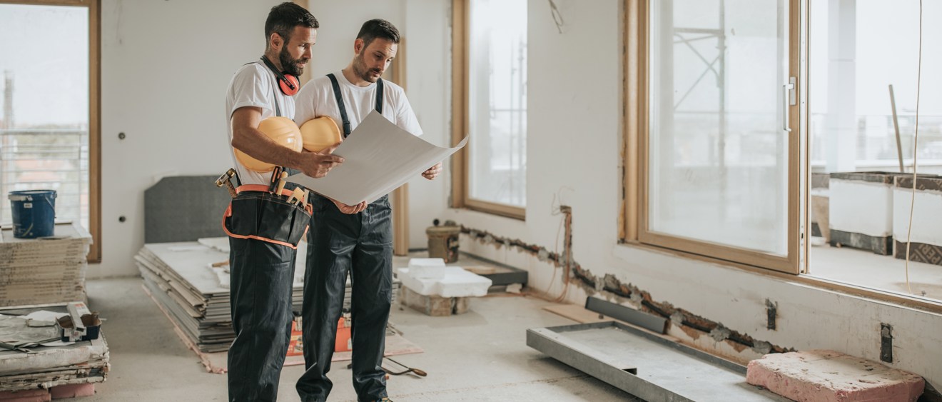 Helbild på två personer som står i en byggnad som verkar vara under konstruktion. De har byggarbetskläder på sig och håller i säkerhetshjälmar. De kollar på ett stort papper som verkar vara en ritning.