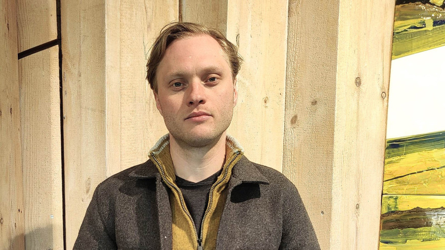 Ossian Hall har blont kortklippt hår som är bakåtkammat. Han har lite skäggstubb och tittar allvarligt in i kameran. Han är klädd i en mörkbrun jacka och bär en ljusbrun tröja med dragkedja under jackan. Han står emot en vägg med bara träplankor.