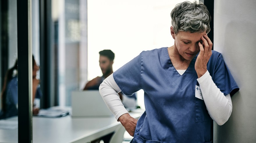 Kvinna i sjukvården vilar huvudet i händerna och lutar sig mot en vägg