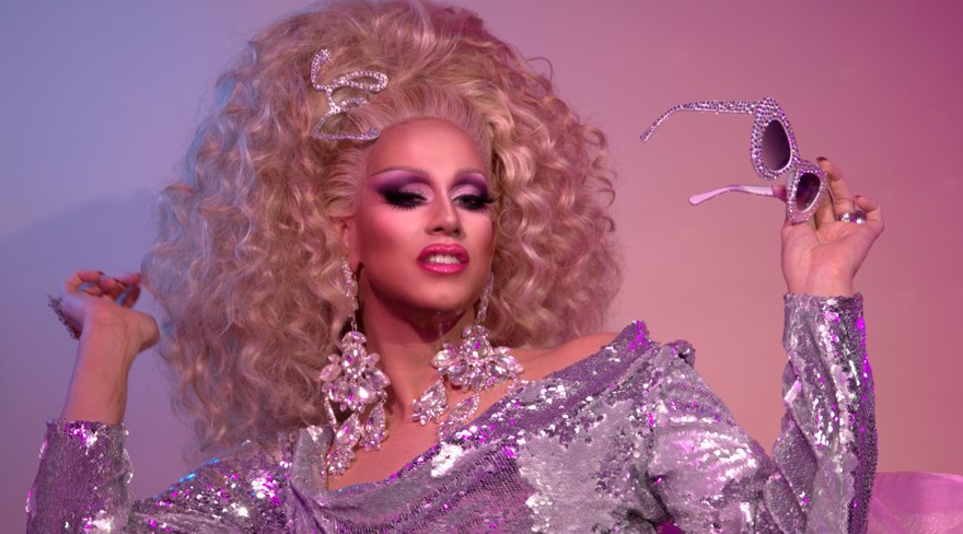 Elecktra sitter bakåtlutad, klädd i en silverglittrig klänning. Elecktra har ett stort blont hår, bär stora glittriga örhängen och håller ett par solglasögon i handen.