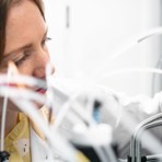 En kvinna står i ett labb, hon bär en vit rock, det bruna håret är uppsatt, blicken är riktad mot en genomskinlig flaska samtidigt som hon skruvar av locket med ena handen som är täckt med en orange plasthandske.