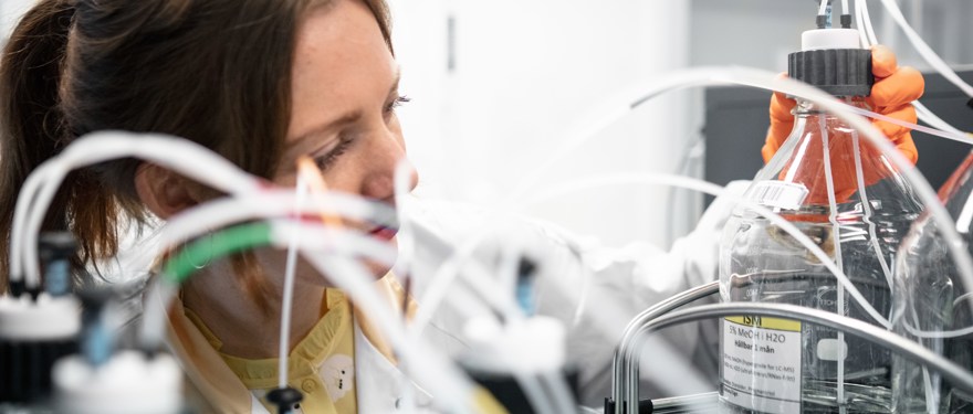 En kvinna står i ett labb, hon bär en vit rock, det bruna håret är uppsatt, blicken är riktad mot en genomskinlig flaska samtidigt som hon skruvar av locket med ena handen som är täckt med en orange plasthandske.