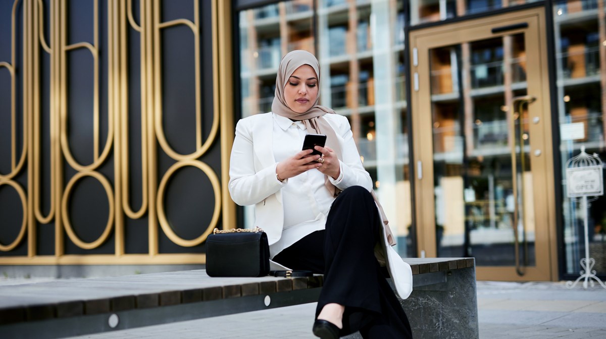 En kvinna sitter på en bänk och tittar i sin mobil.