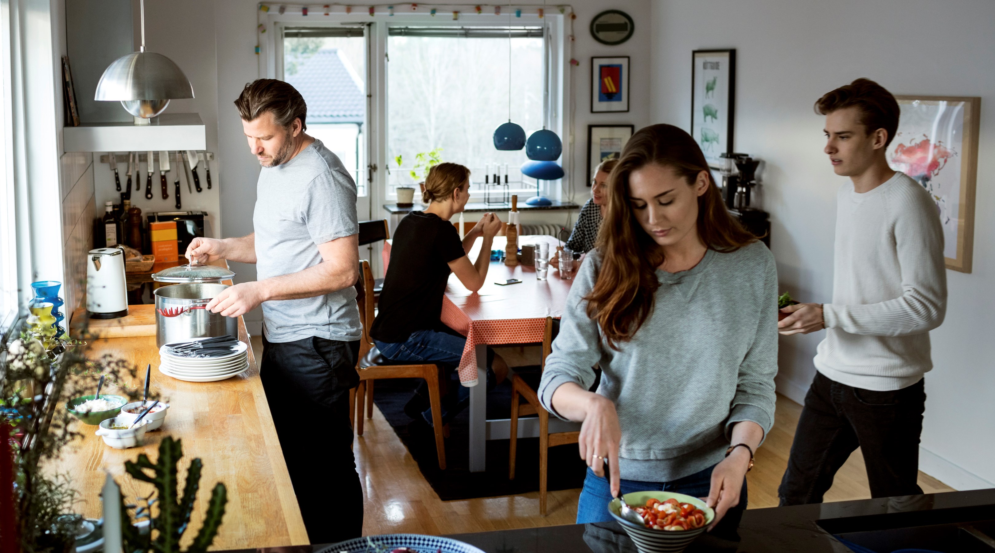 En man står och tar mat ur en gryta och en kvinna förbereder en sallad. Flera människor sitter redan till bords.