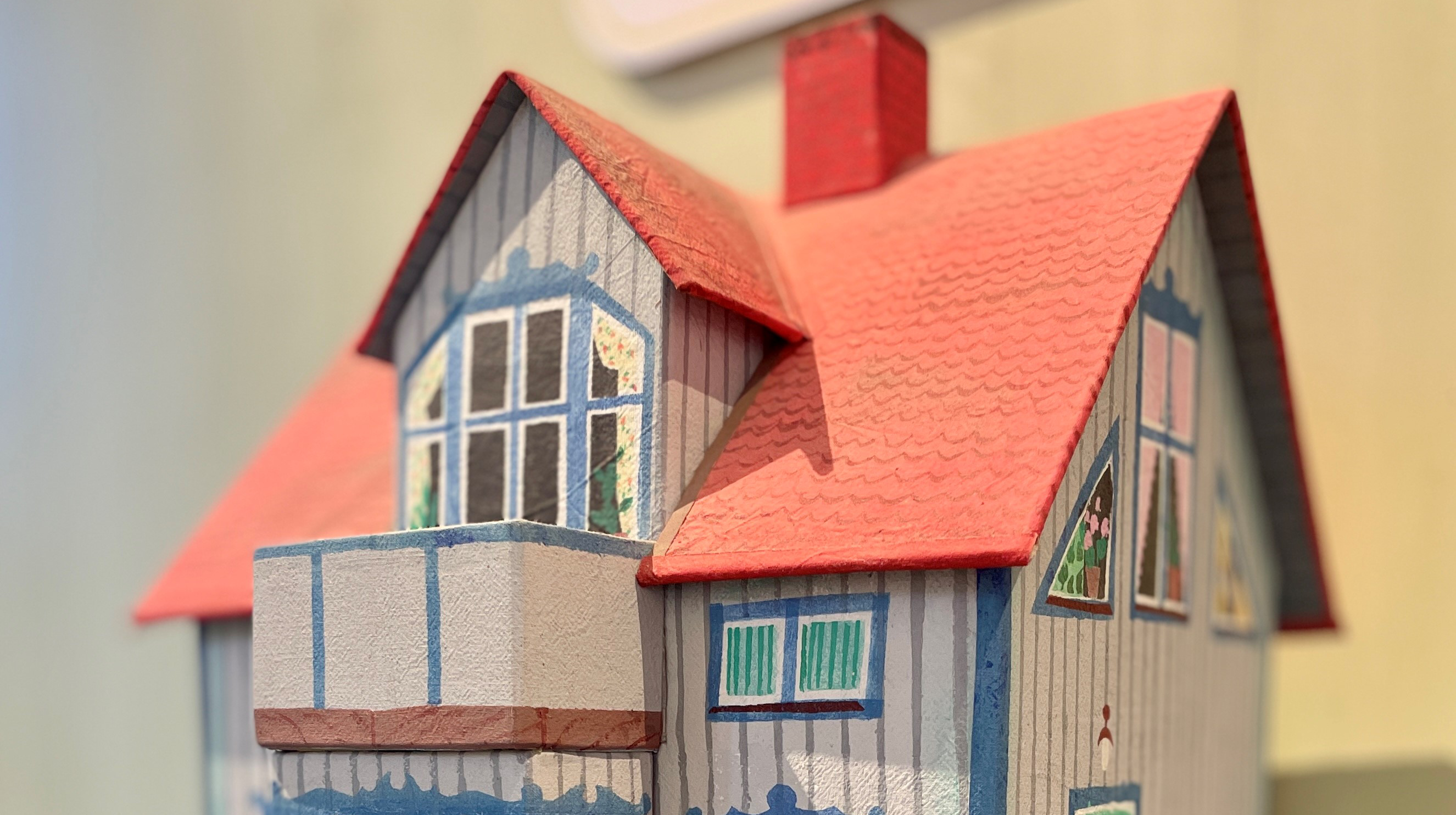 En modell av en villa med rött tak och blågrå panel målat, i fönstren målade gardiner och blommor.