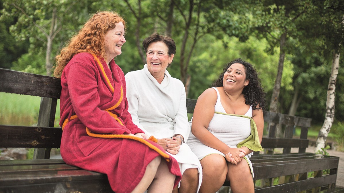Tre kvinnor sitter på en bänk. De ler mot varandra. De har badrockar  på sig.