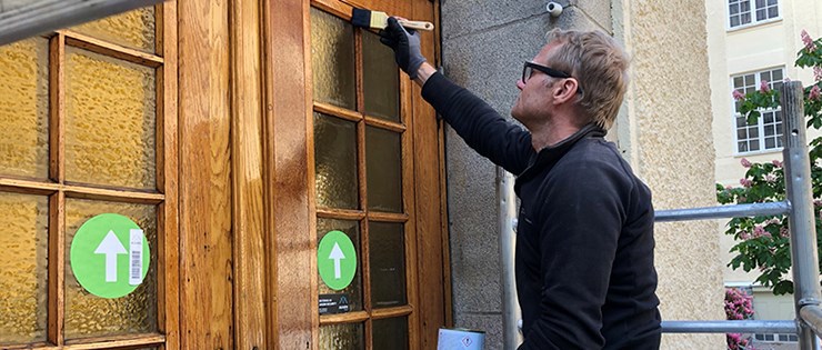 En person i mörkblå arbetskläder sitter på en byggnadsställning och fernissar en trädörr med gula glas. Hen håller en pensel i höger hand och en burk färg i vänster.