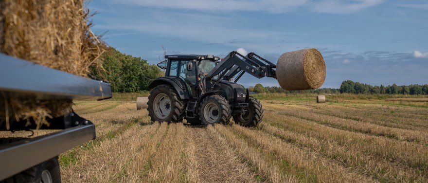 Traktor med lastare lyfter upp halmbal på skördat fält med balvagn i förgrunden