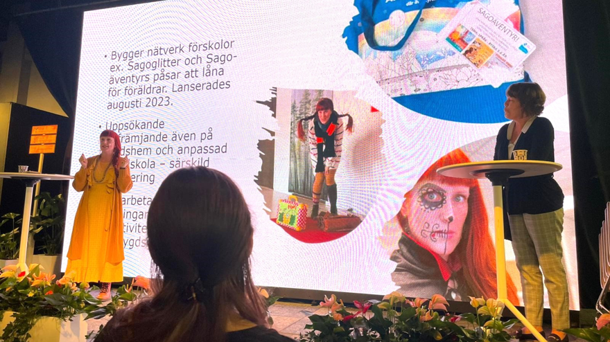 Två kvinnor står på en scen, en är klädd i en gul klänning, den andra bär rutiga byxor och en svart tröja. De pratar båda två, bakom dem syns en stor skärm med text och bilder. I mitten av bilden syns bakhuvudet på en kvinna i den lyssnande publiken.