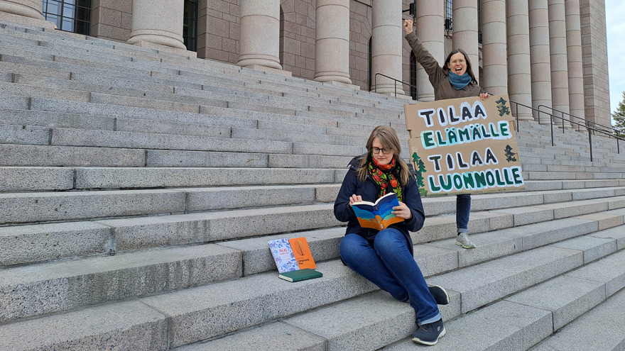 Liisa Uimonen och Johanna Juvonen befinner sig utomhus på en stor stentrappa till en stor byggnad. Liisa sitter och bläddrar i böcker medan Johanna står med ett plakat och håller en knuten näve i luften.