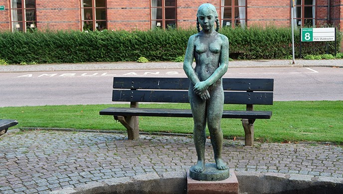 En skulptur av en ung flicka framför en bänk. I fonden ett tegelhus.