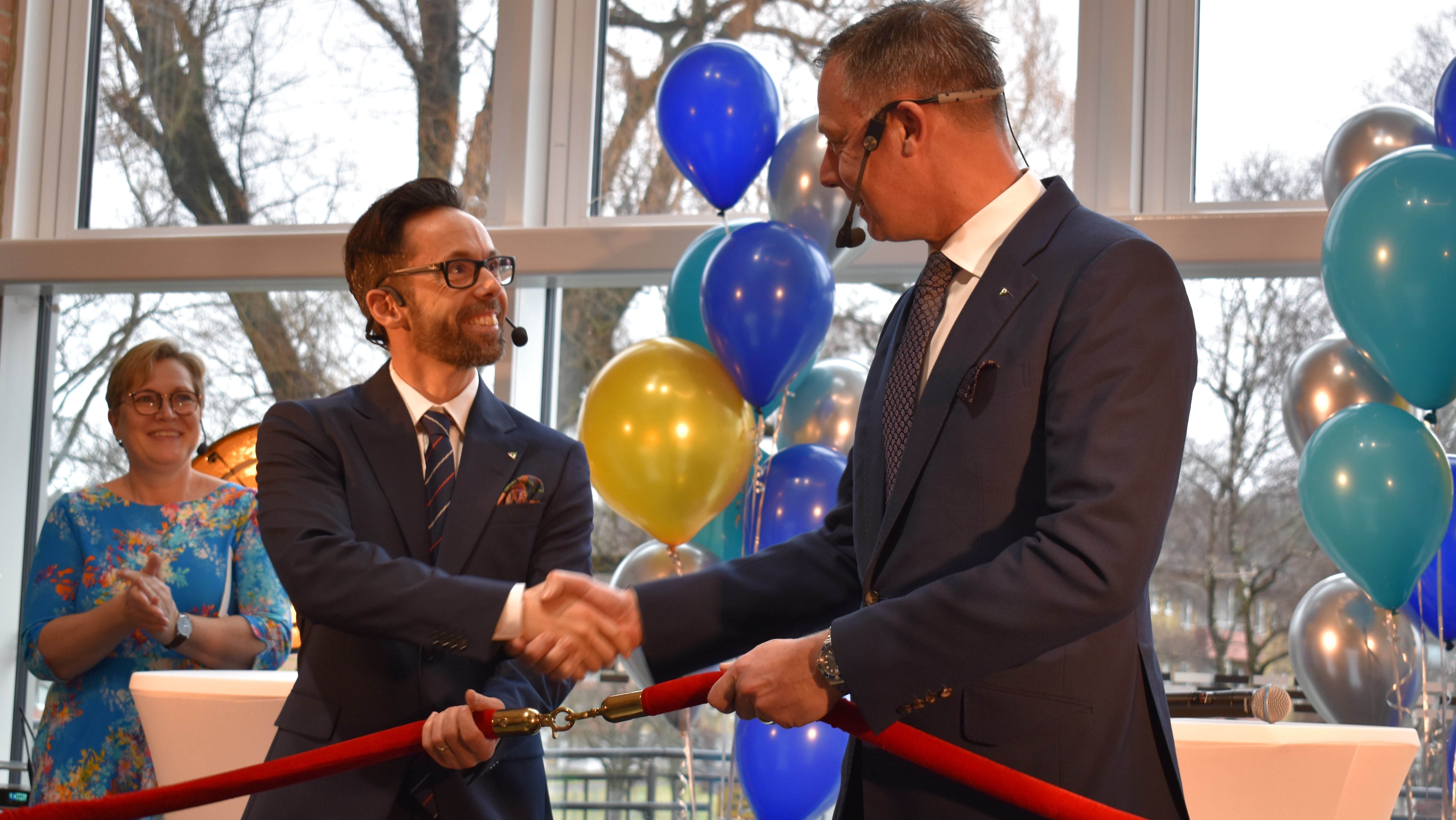 Män i kostym, skakar hand, ler och håller i ett sammanlänkat band. Ballonger och en glad kvinna i bakgrunden.