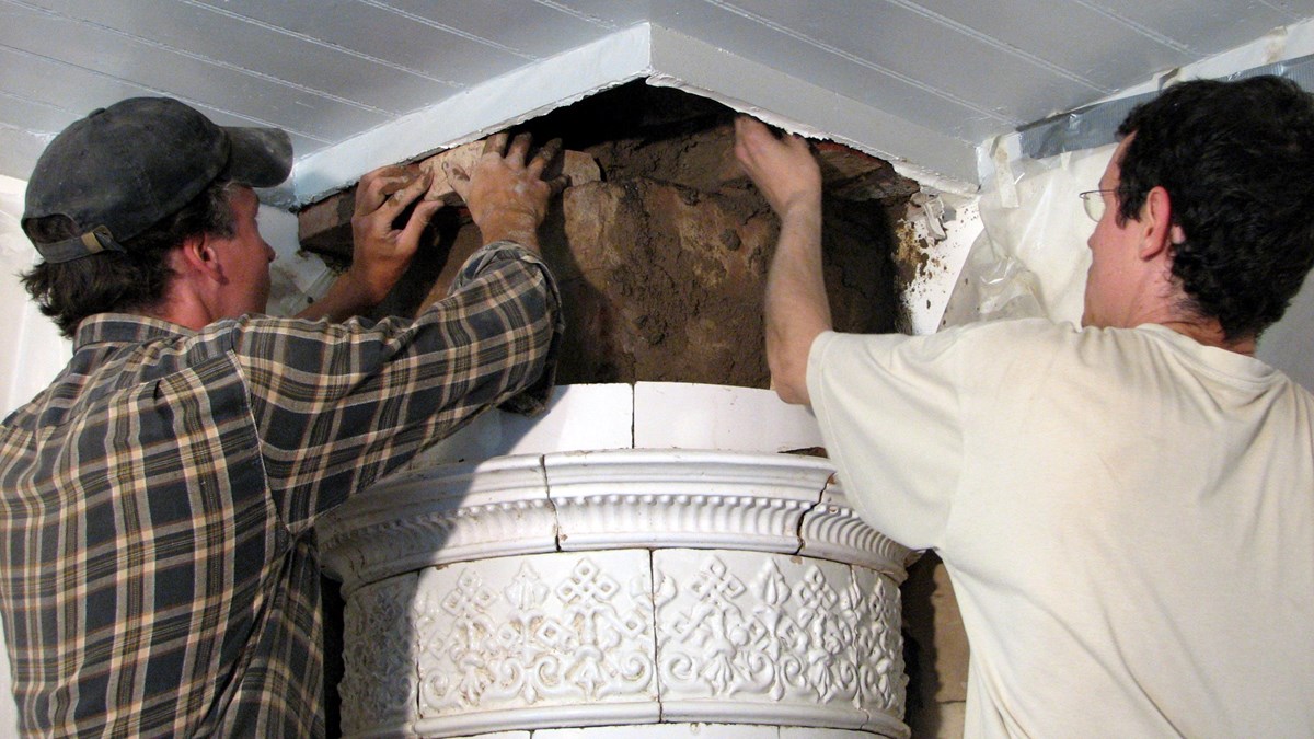 Två personer renoverar en vit kakelugn, nära taket
