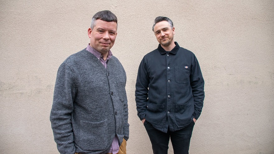 Johannes Jensen har kortklippt gråsprängt hår och är klädd i en grå kofta och en mönstrad skjorta. Kristian Schultz har gråsprångt bakåtkammat hår och skägg och är klädd i en svart skjorta. Bakom dem syns en beige rappad vägg.