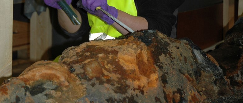 En rostig järnklump ligegr på en yta. Ett par händer i lila latexhandskar knackar försiktigt bort krustan för att få fram det som finns i den rostiga järnklumpen.
