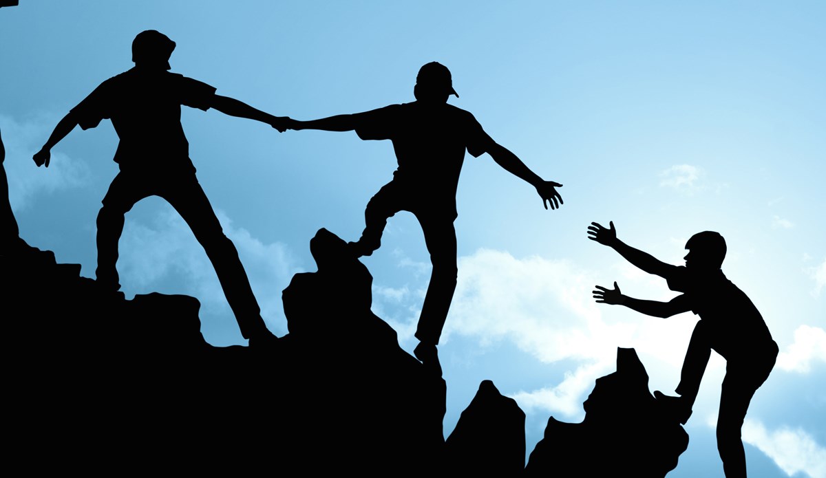 Bilden visar tre mänskliga siluett mot en blå himmel. Personerna  hjälper varandra uppför ett steningt berg.