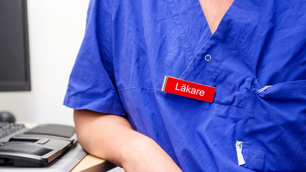 Närbild på läkares blå arbetskläder med skylten "läkare" på tröjan