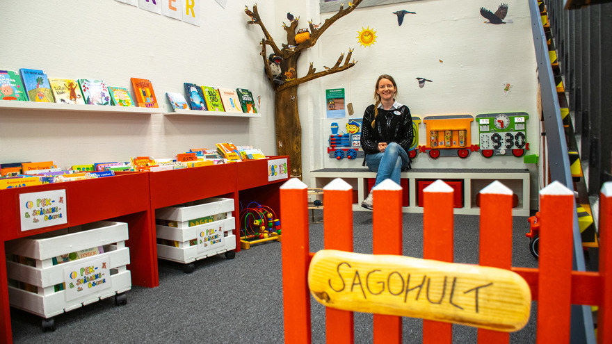 Erika Ribacke Sandberg sitter i småbarnshörnan Sagohult på Melleruds bibliotek. Boktrågen är röda liksom en bit staket som finns i förgrunden. Erika har långt blont hår i en fläta, är kläd i jeans och en svart tröja. Hon sitter på en bänk mot väggen där också ett stiliserat träd finns som prydnad.