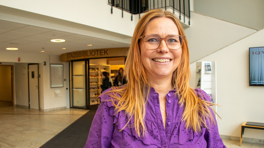 Maria Nordin är i medelåldern, har långt blont, rakt hår och bär glasögon. Hon är klädd i en lila blus och bakom henne skymtar en entré med texten STADSBIBLIOTEK.