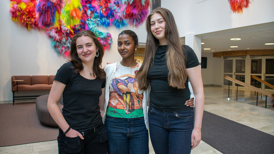 Alvina Birgersson, Fahmo Mohamed och Hannah Evensen är alla i 20-årsåldern. Alvina och Hannah står på varsin sida om Fahmo och de håller varandra om ryggen. De ler mot kameran, i bakgrunden syns ett färgglatt konstverk på en vägg, en soffa och en trappa.