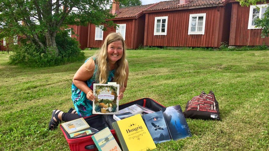 Anna Hällgren har långt ljust hår och är klädd i en ärmlös mönstrad klänning. Hon sitter framför en boklåda utomhus.