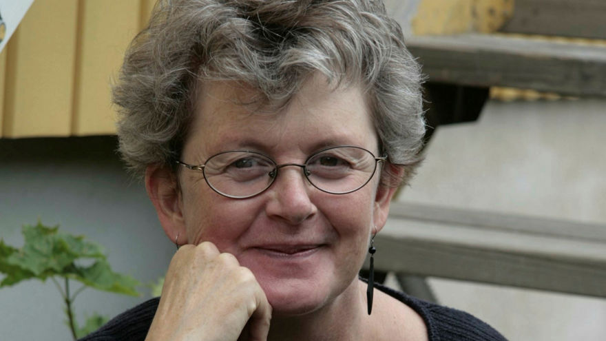 Lotta Olsson har gråsprängt kort lockigt hår. Hon bär ett par ovala glasögon med tunna bågar. Hon tittar rakt in i kameran och ler med stängd mun.