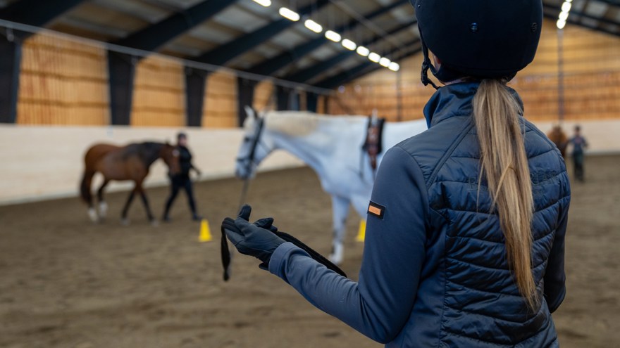 Elev med ridhjälm står framför vit häst i ridhus
