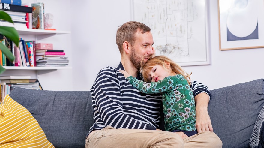 Fotografi taget framfrån. En pappa håller om sitt barn. De sitter i en grå soffa.