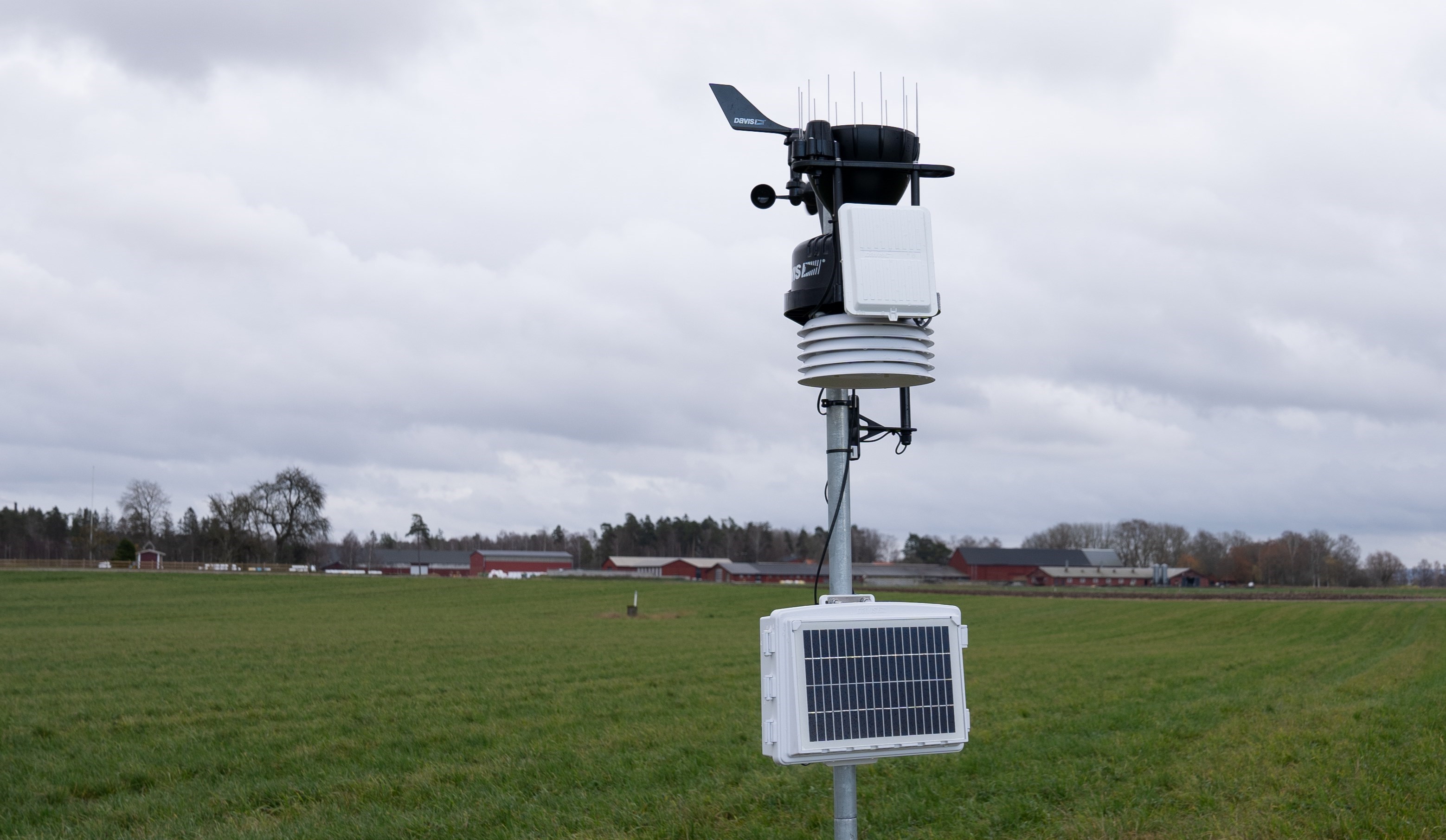 En väderstation av sammankopplade sensorer står på ett fält. I bakgrunden syns Naturbruksskolan Uddetorp.
