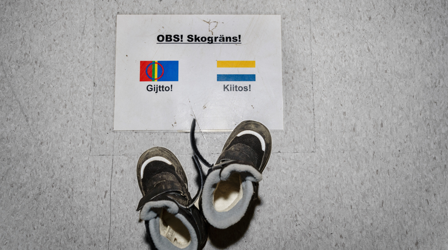 Skylt på golvet med texten Skogräns. Vid skylten står ett par barnskor. På skylten står det tack på både samiska och meänkieli.