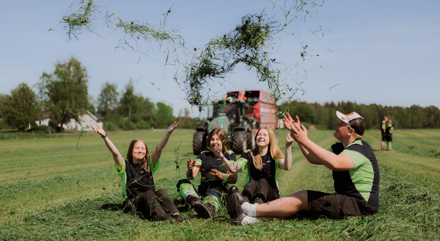 Fyra elever sitter på marken och kastar upp nyslaget gräs i luften