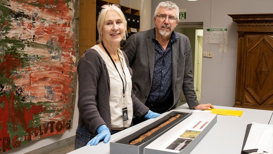 En kvinna med ljust hår står bredvid en en gråhårig man med glasögon. Kvinnan har blå latexhandskar på sig. På bordet intill dem ligger en pappask med ett formskuret underlägg där ett gammalt, rostigt svärd ligger. Båda personerna tittar in i kameran och ser glada ut.