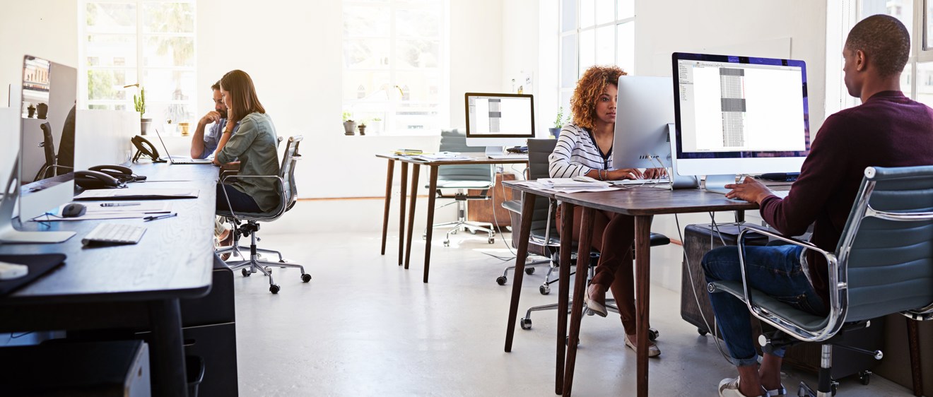 Helbild som visar ett öppet kontorslandskap där flera personer sitter och arbetar framför en dator med stor bildskärm.