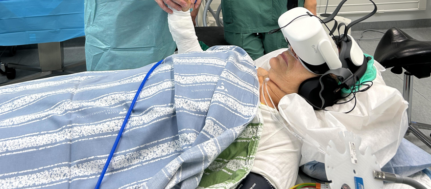 läkare och sjuksköterska står bakom säng som patient ligger i. Patienten har på sig VR-glasögon.