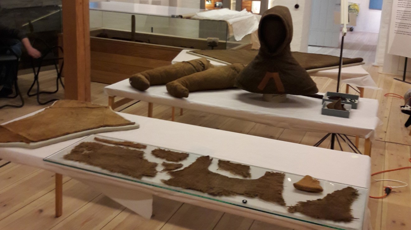 I en lokal står ett par bord med vita bomullsöverdarg uppställda. På det främre bordet syns bruna tygstycken. På det bakre skymtar en struthätta samt ett par hosor. Alla textilierna är bruna.