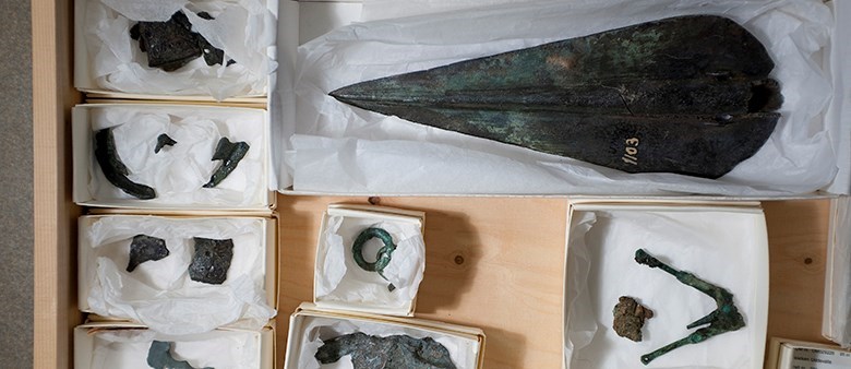 Arkeologiska fynd i metall, bland annat en spjutspets, ligger packade i silkespapper i vita pappaskar i en trälåda.