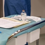 En stålvagn på hjul står i ett rum på ett sjukhus. Vagnen är täckt med blått papper, på vagnen ligger det A4 papper med text, en bläckpenna, en genomskinlig glasflaska med etikett och bredvid ligger det sprutor som är inplastade. I bakgrunden syns en person i blå läkarrock.