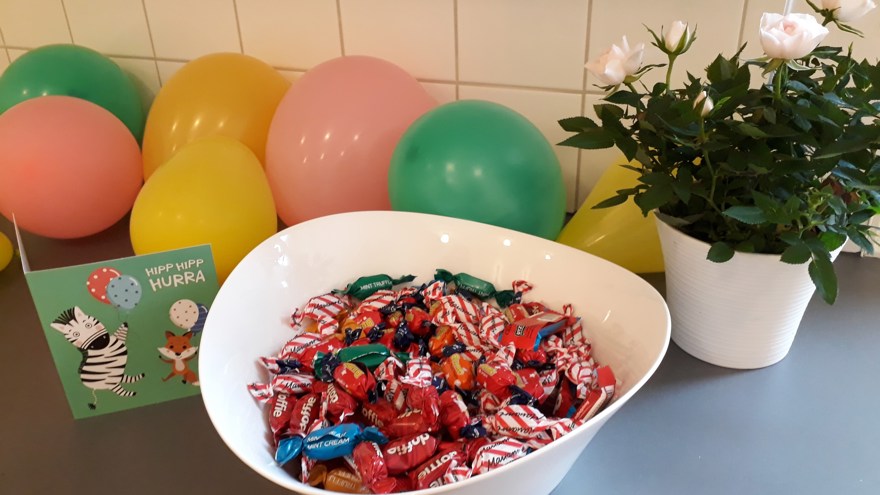 Bild på ett bord med ballonger och en skål godis, en blomma och ett grattiskort