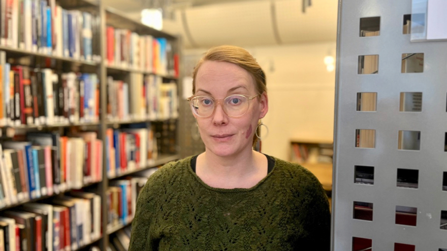 Eeva Östberg har blont hår som är uppsatt. Hon har glasögon och bär en grön stickad tröja. På båda sidor om henne finns bokhyllor fyllda med böcker.