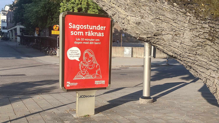 En gata i stan. På en inglasad skylt står texten "Sagostunder som räknas - Läs tio minuter om dagen med ditt barn!".