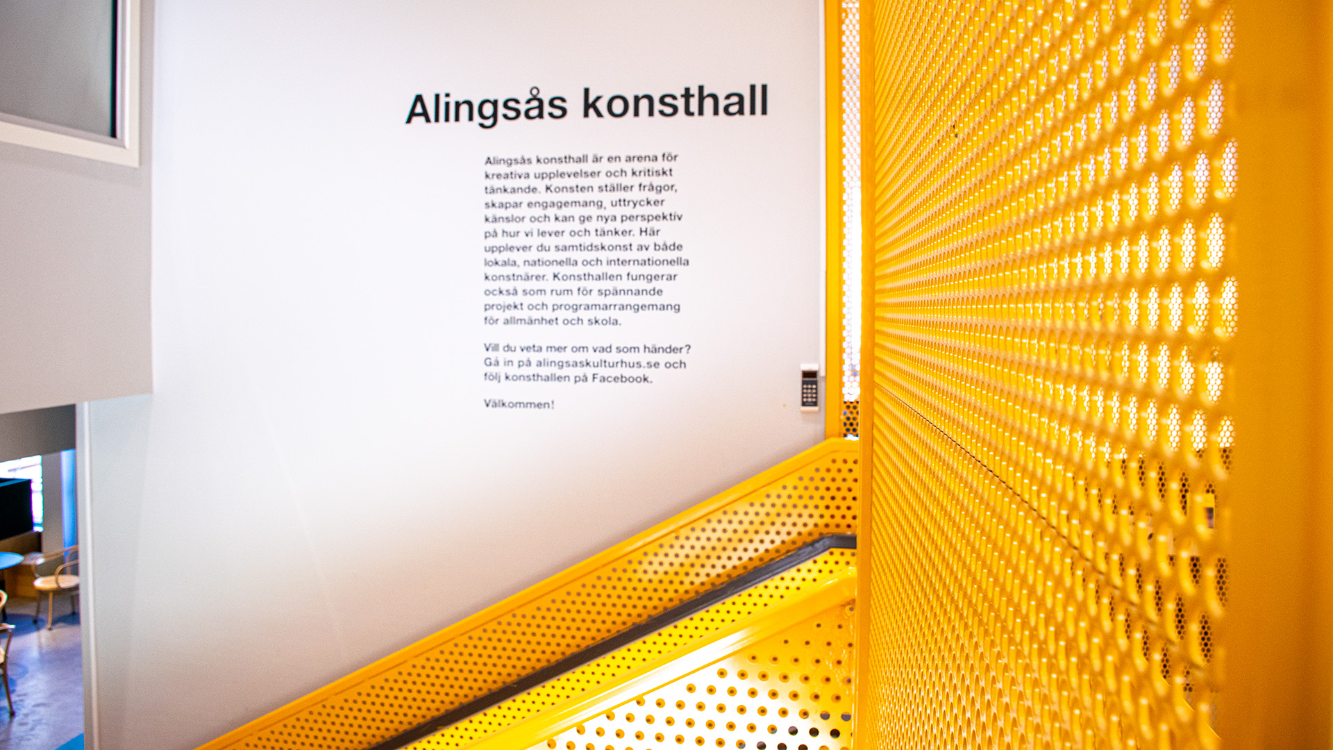 På en vit vägg står det Alingsås konsthall och en text under rubriken. Texten står i trappan på väg upp till konsthallen, trapphuset domineras av de gula perforerade metallskydden som omger trappstegen i trappan. 