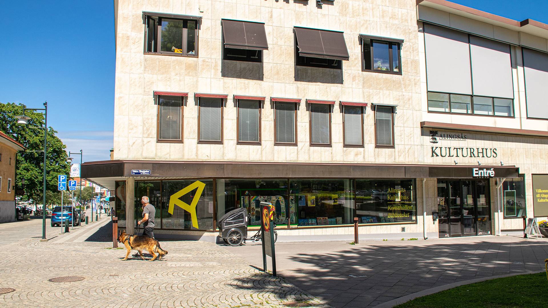 Kulturhuset i Alingsås. En byggnad i tre våningar med beige fasad. LÄngst ner ligger biblioteket och mot gatan vetter stora glasfönster. På fasaden står det Alingsås Kulturhus och över dörren står det Entré. En man med en schäfer går förbi, solen skiner och längre bort i bild står en parkerad blå bil och gröna stora träd.