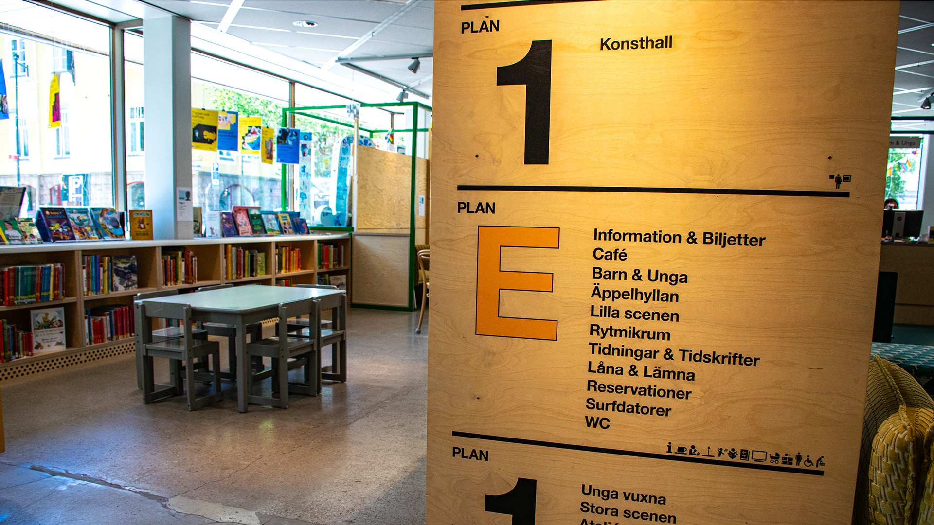 En informationsskylt visar vad som finns på Kulturhusets olika våningar. Skylten är i trä, den står på golvet i bibliotekets barnavdelning. Fönstren i lokalen vetter ut mot gatan, här finns låga bokhyllor och ett lågt bord med fyra stolar för barn.  