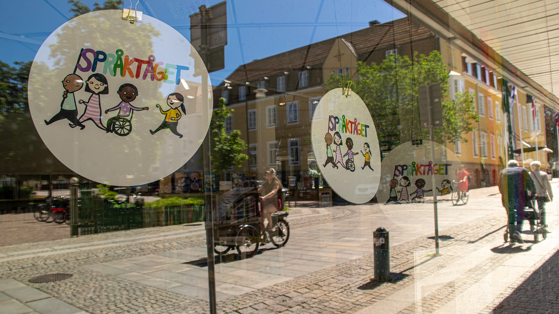 Alingsås speglas i bibliotekets skyltfönster. Bilden är tagen utifrån, solen skiner. I fönstren hänger runda skyltar med texten Språktåget. I speglingen i fönstren cyklar en tjej förbi på en lådcykel. Ett gult äldre stenhus speglas i glaset och ett äldre par vandrar på gatan utanför. Mannen går med rollator.  