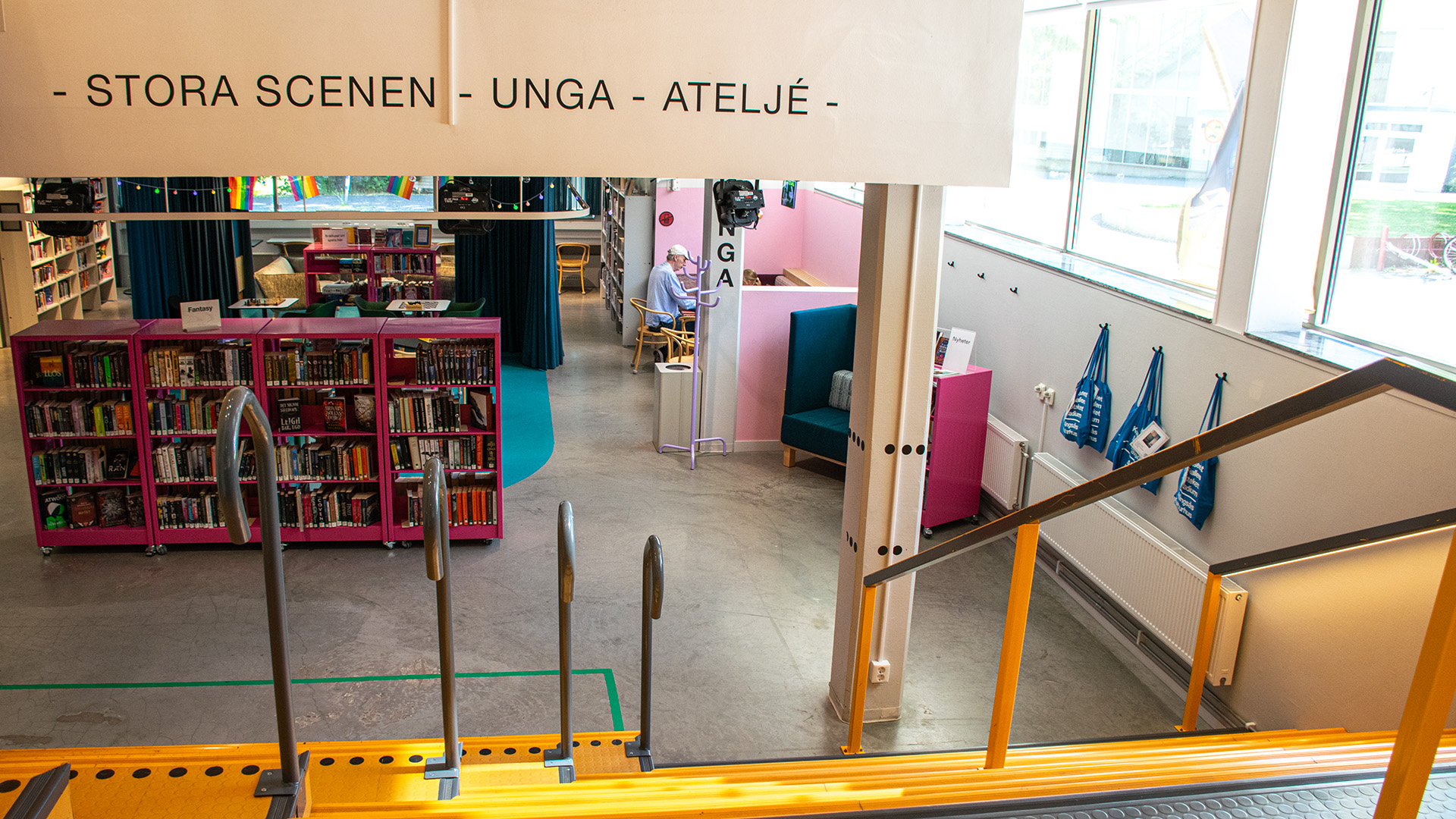En gul trappa leder ner till Kulturhusets lägsta våning. På en skylt i taket står det: STORA SCENEN - UNGA - ATELJÉ. En bokhylla rakt fram är målad i en knallrosa färg, en blå matta ligger på golvet bakom den och där finns två stora blå draperier som hänger från taket. En äldre man med en vit keps sitter vid en dator vid ett fönster. . 