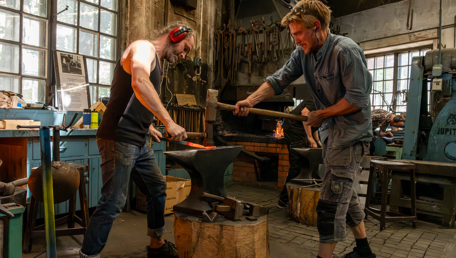 Två smeder arbetar ut ett glödgat styck järn med hjälp av hammare och slägga mot ett städ.