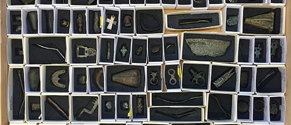Bilden visar arkeologiska föremål. Alla ligger i varsin vit pappask på ett mjukt, mörkgrått underlag där en fördjupning är utskuren som passar respektive föremål.