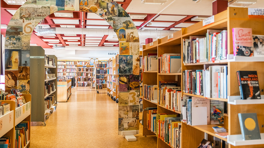 Biblioteket i Hyltebruk. Bokhyllor står i raka rader, ett rakt gångstråk leder framåt, längst bort finns en vägg med fler bokhyllor fyllda med böcker. Till höger finns boktråg med bilderböcker, på övriga hyllor finns skyltar som bland annat Världslitteratur, Fakta och Spänning. En byggd båge spänner över gångstråket, den är täckt med ett kollage av bilder från bilderböcker.