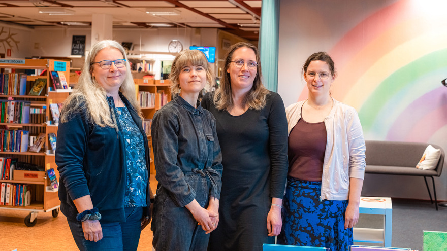 Fyra kvinnor står bredvid varandra i ett bibliotek. De står uppställda för fotografering, alla tittar mot fotografen. Två av kvinnorna är i medelåldern, två av dem något yngre. Bakom finns bokhyllor med böcker, en vägg med en målad regnbåge och en grå soffa. På väggen i bakgrunden hänger en klocka som visar tiden.
