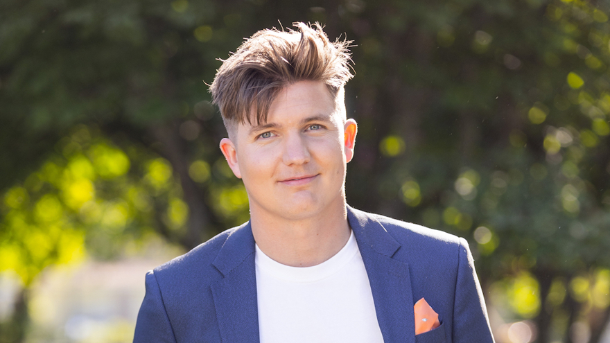 Daniel Norberg står utomhus, solen skiner. Han har kortklippt ljusbrunt hår och ljusa ögon. Han är klädd i en blå kavaj och en vit t-shirt. Han är i 35-årsåldern.